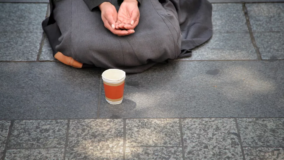 A woman with hands folded and a cup in front of her, on the sidewalk