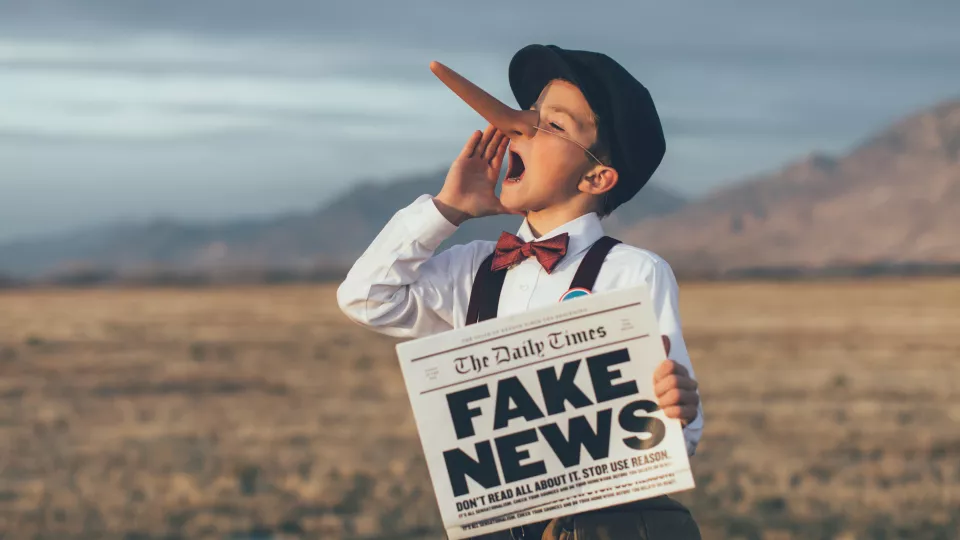 A child with a fake, long nose holding a newspaper that says 'fake news'. Photo.