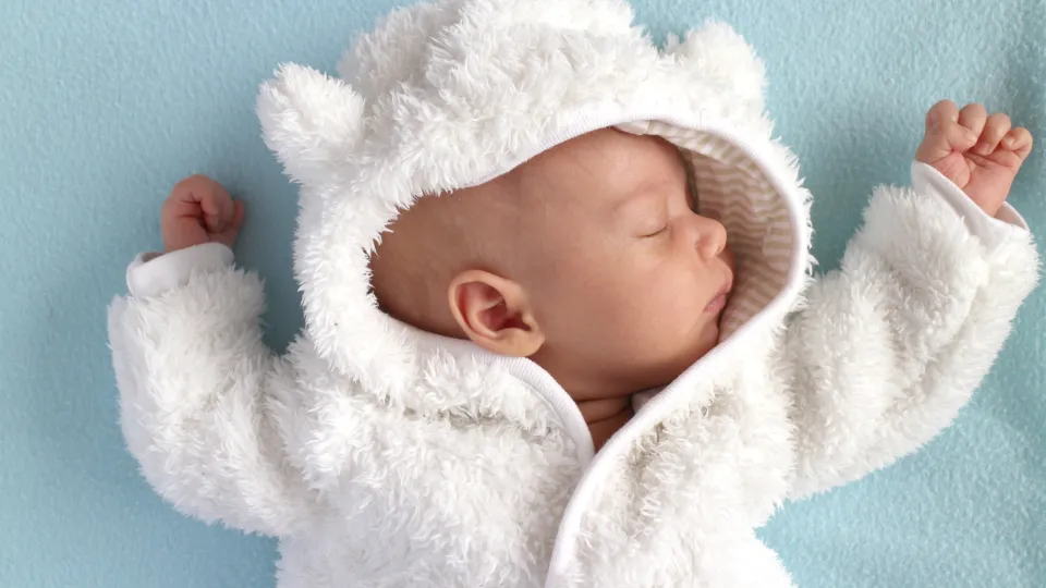 Sleeping cute baby in a white onepiece against blue background, photography. Photo: iStock