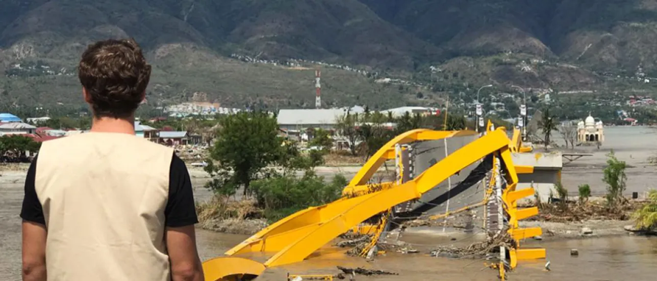 A person stands with their back to the camera, looking out over a disaster-stricken area.