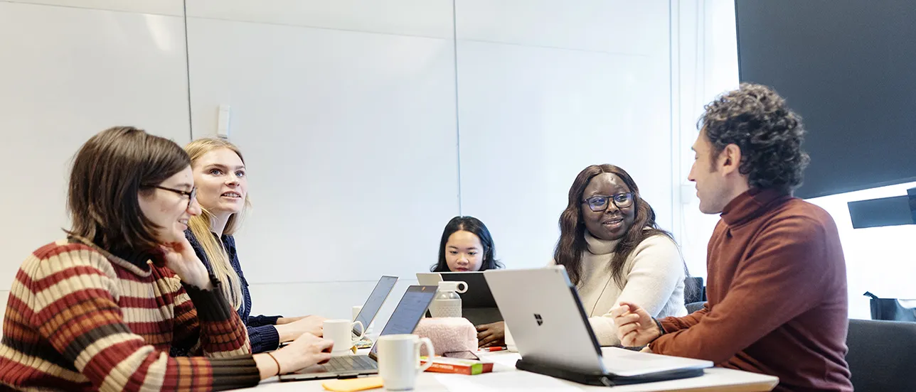Five students interacting in a classroom with their laptops in front of them. Photo.