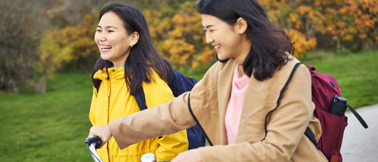 Two students in an autumn scene, one of them leading a bicycle. Photo.