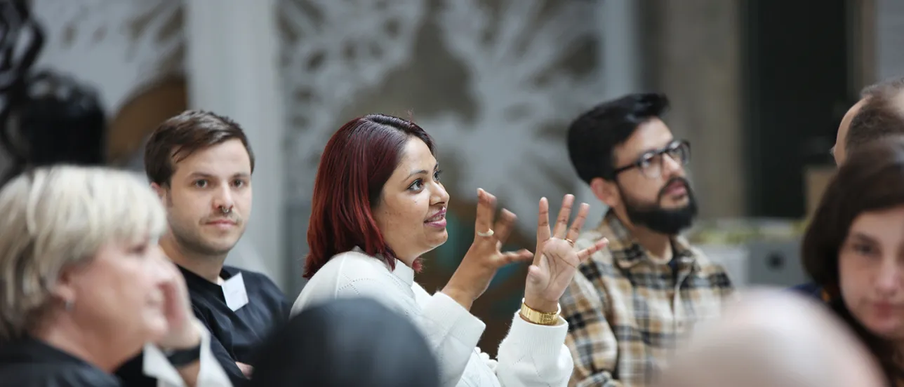 A group of people seated around a table engaged in discussion, with one person in the center speaking and gesturing. Photo.