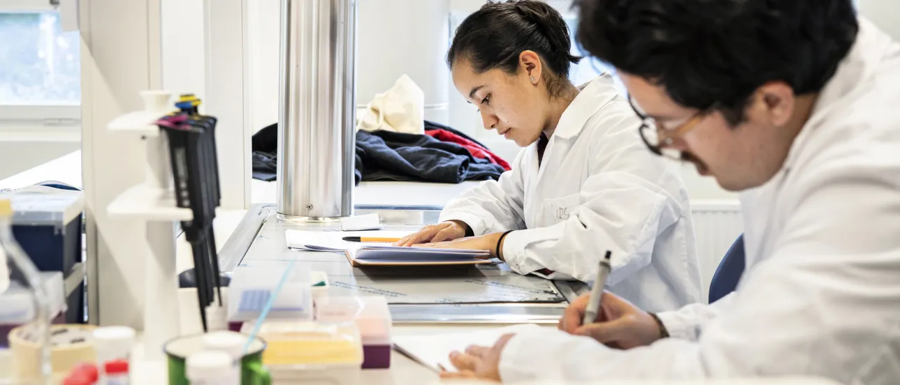 Students in lab coats are writing notes.