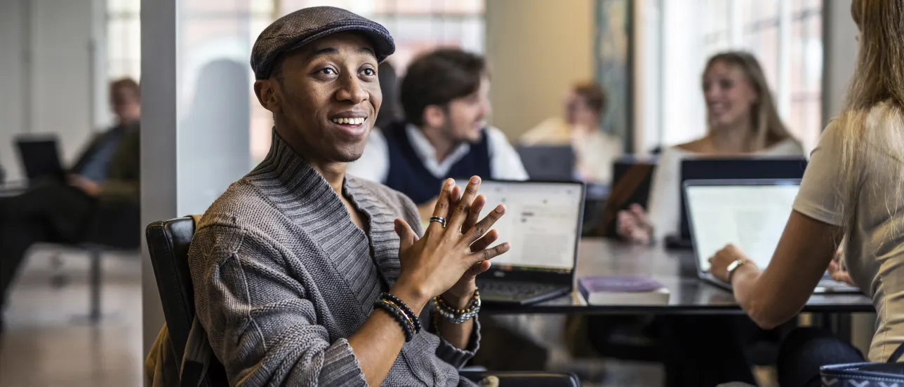 A student sits at a study table in the hallway and looks out. More students are studying together in the background. Photo.