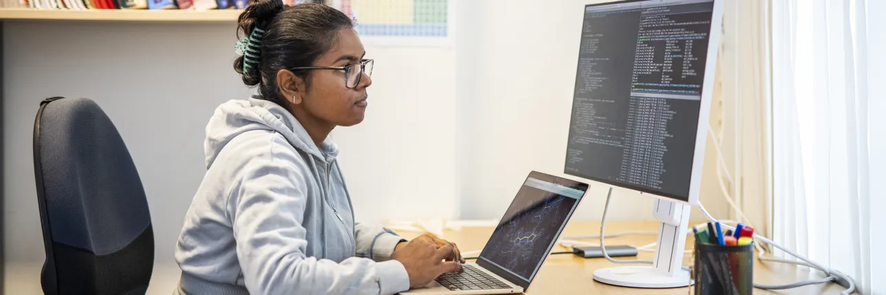 Person working at a desk on a laptop next to a large external monitor displaying code. Photo.