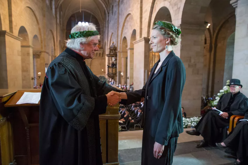 A doctoral candidate with a laurel wreath receiving a diploma with a handshake