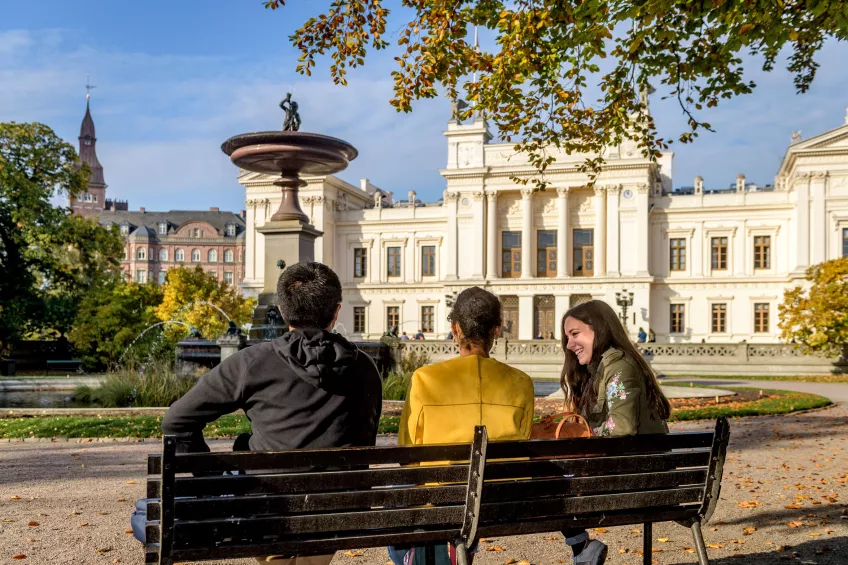 Three students talking on a bench in front of the fountain and the Main University Building