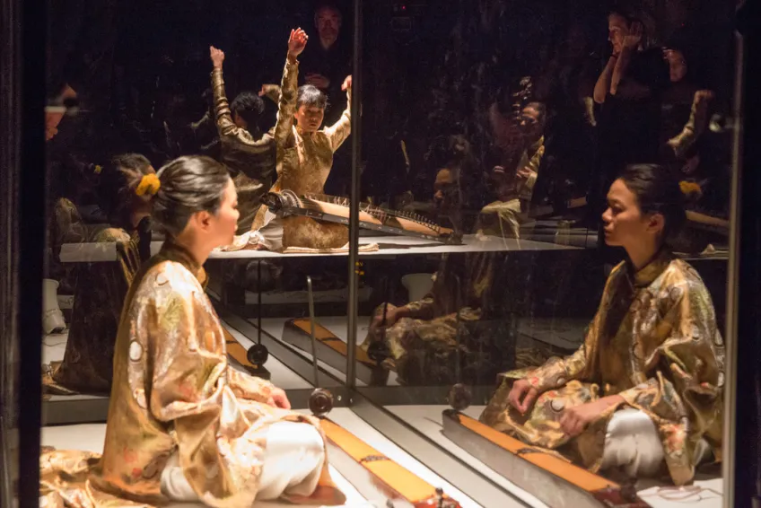 A performance with someone looking at their reflection in a window and someone playing some sort of instrument