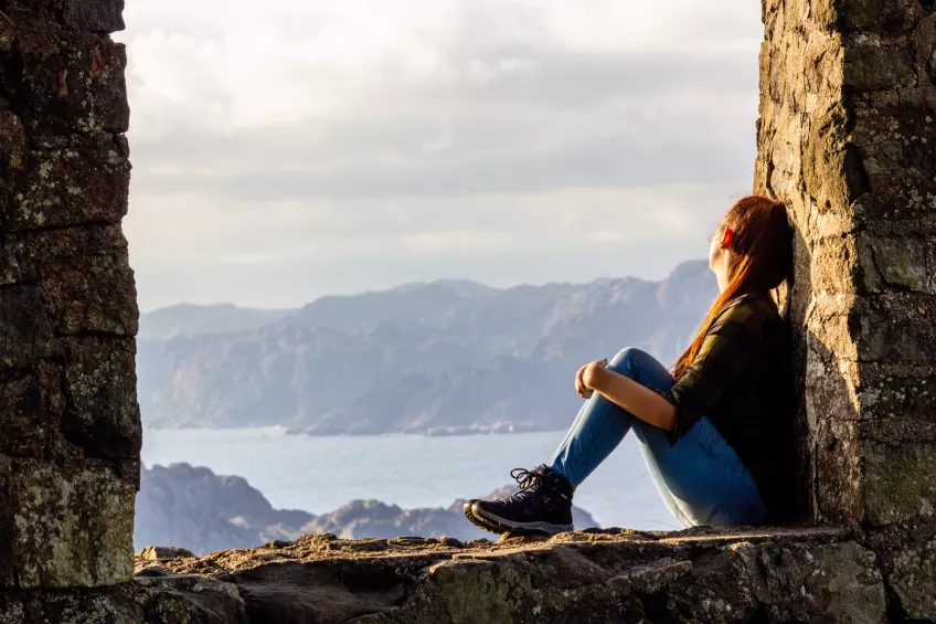 Someone sitting in between two stone walls looking out over a Norwegian landscape with sea and mountains. Photo. 