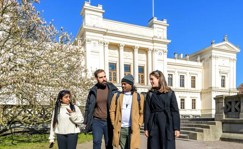 Four students walking in front of the main university building in Lund. Photo.